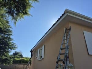 Ouvrier sur une échelle installant ou nettoyant des gouttières sur une maison orange sous un ciel bleu.