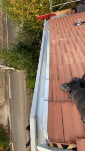 Couvreur posant une gouttière en zinc sur une toiture en tuiles rouges avec vue plongeante sur la rue.