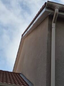Façade en crépi beige avec gouttière blanche et toiture de tuiles rouges sous un ciel nuageux.