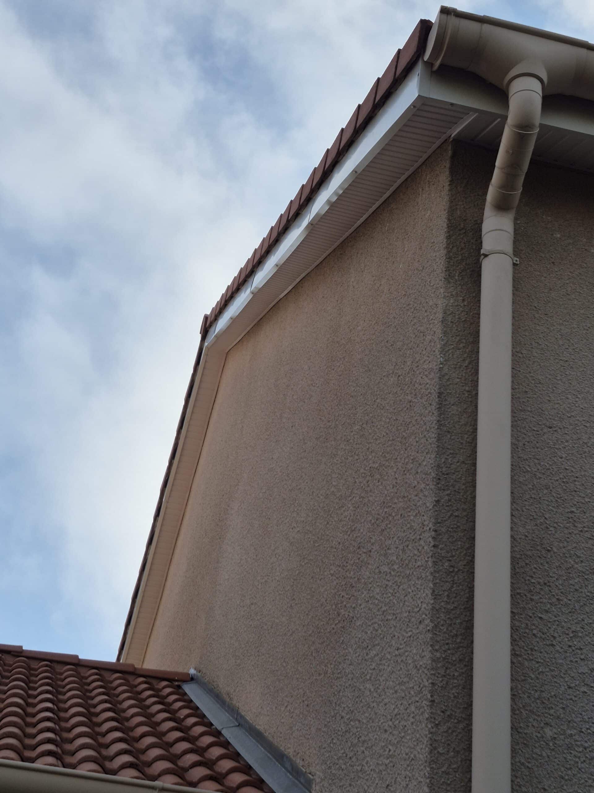 Façade en crépi beige avec gouttière blanche et toiture de tuiles rouges sous un ciel nuageux.