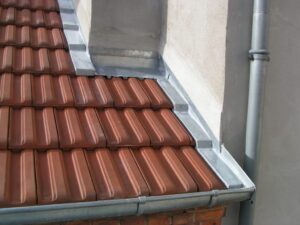 Étanchéité de toiture avec tuiles rouges, solin et gouttière en zinc contre un mur extérieur.
